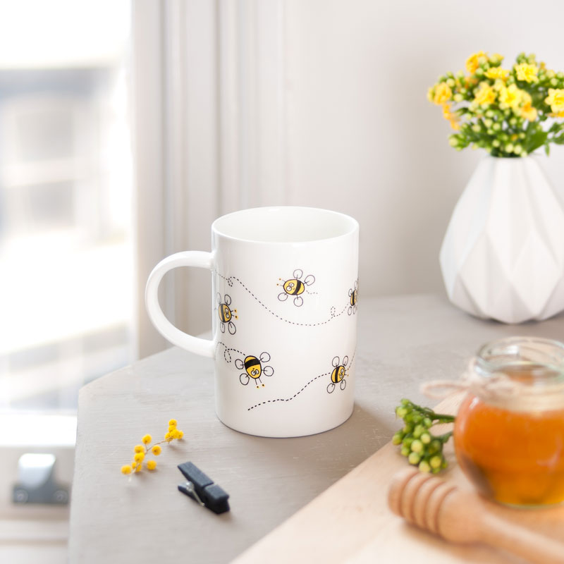 The reverse of a white mug with bee design on a table with flowers and honeycomb