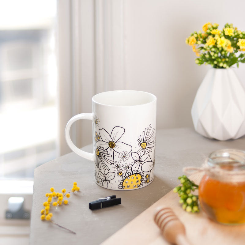 White mug with floral and bee design on a table with a vase of flowers and a honey pot.